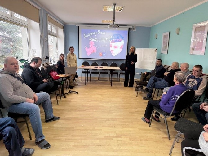 W sali spotkań osoby siedzą w kręgu, patrząc na ekran z prezentacją o Światowym Dniu Walki z Depresją. Dwie osoby stoją przy ekranie, a pozostali słuchają na ustawionych wokół krzesłach.