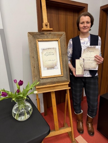 Kobieta stoi obok drewnianej sztalugi z dużą ramą, w której znajduje się ozdobny dokument. W rękach trzyma podobny dokument i nagrodę w pudełku. Obok stoi stół z wazonem fioletowych tulipanów.