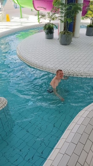 Mężczyzna stoi w płytkiej części basenu, otoczona jasną, turkusową wodą. Wokół znajdują się zaokrąglone kafelkowe brzegi oraz rośliny w donicach na podwyższonym, białym podeście. W tle widać kolorowe elementy zjeżdżalni.