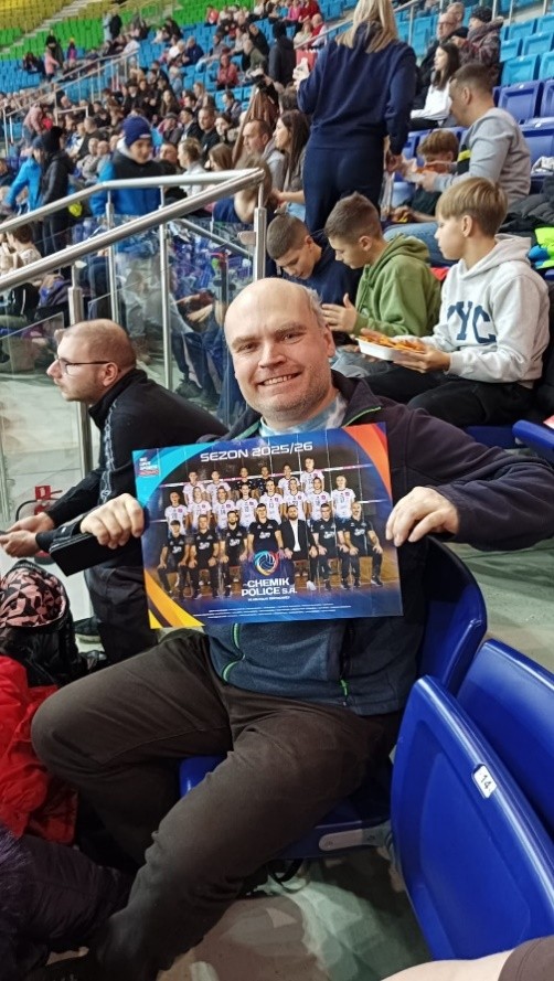 Osoba siedzi na trybunach hali sportowej, trzyma plakat drużyny siatkarskiej z napisem „Sezon 2025/26 Chemik Police S.A.”. W tle znajdują się kibice na kolorowych krzesełkach.