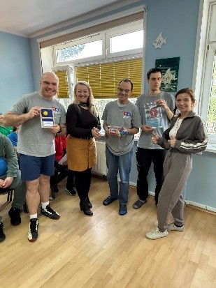 Grupa uśmiechniętych osób stoi w jasnym pomieszczeniu przy oknie. Kilka osób trzyma dyplomy i drobne upominki. 