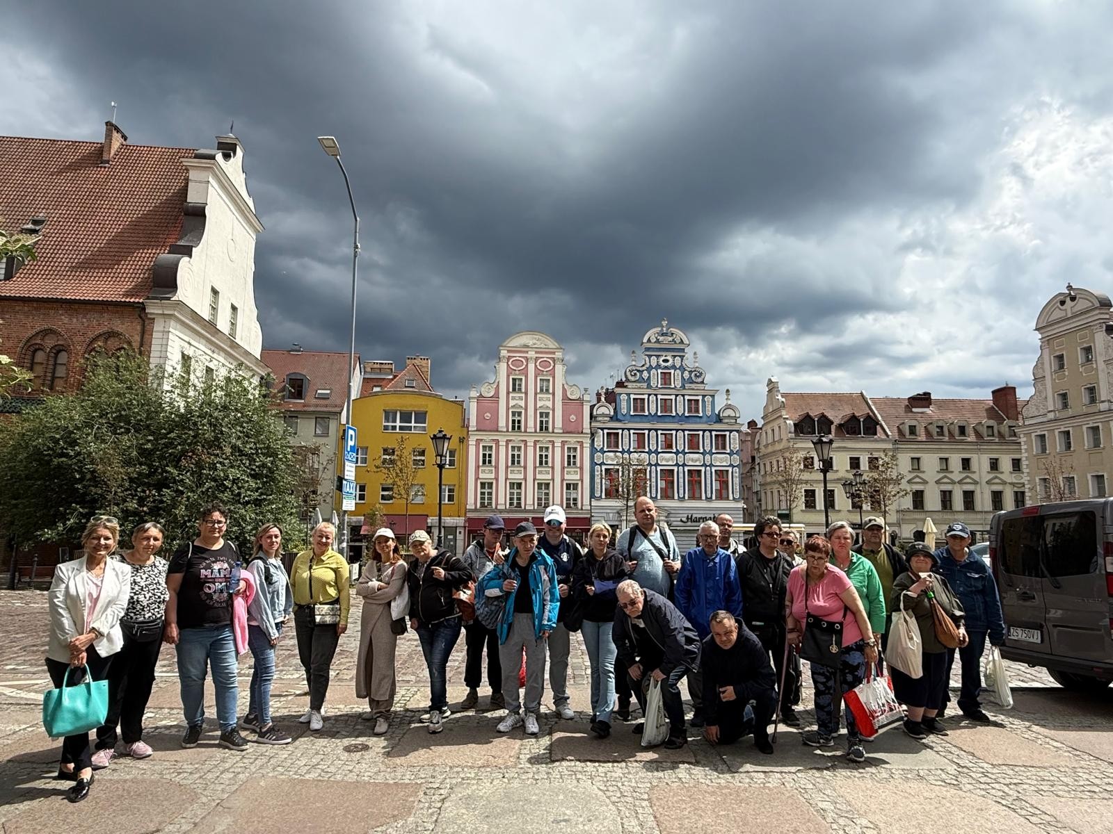 Grupa osób stoi na Starym Mieście i pozuje do zdjęcia. Na niebie ciemnoszare chmury i jednocześnie ulice oświetlone są słońcem.