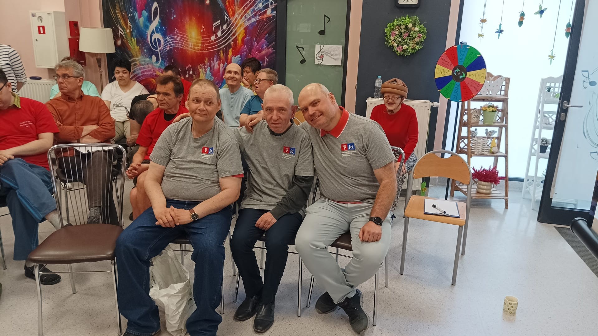 Trzech mężczyzn w szarych podkoszulkach siedzi na krzesłach i uśmiecha się do zdjęcia. W tle wiele innych siedzących osób.