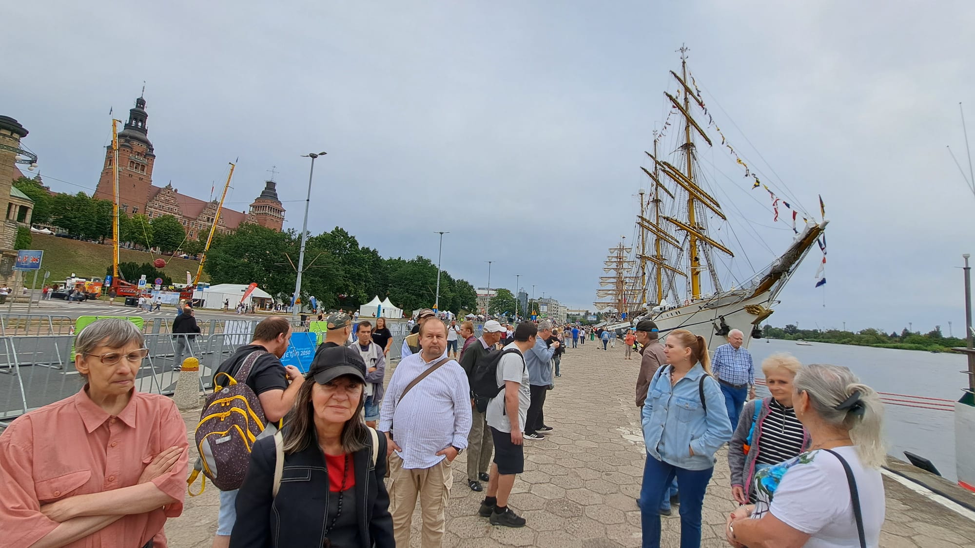 Grupa osób stoi na bulwarach na tle łodzi żaglowych - żaglowców morskich.