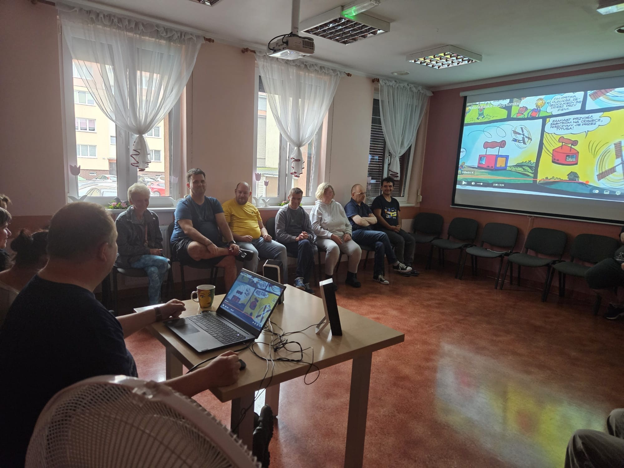 Grupa osób słuch wykładu o komiksach,na ekranie slajd z komiksami.