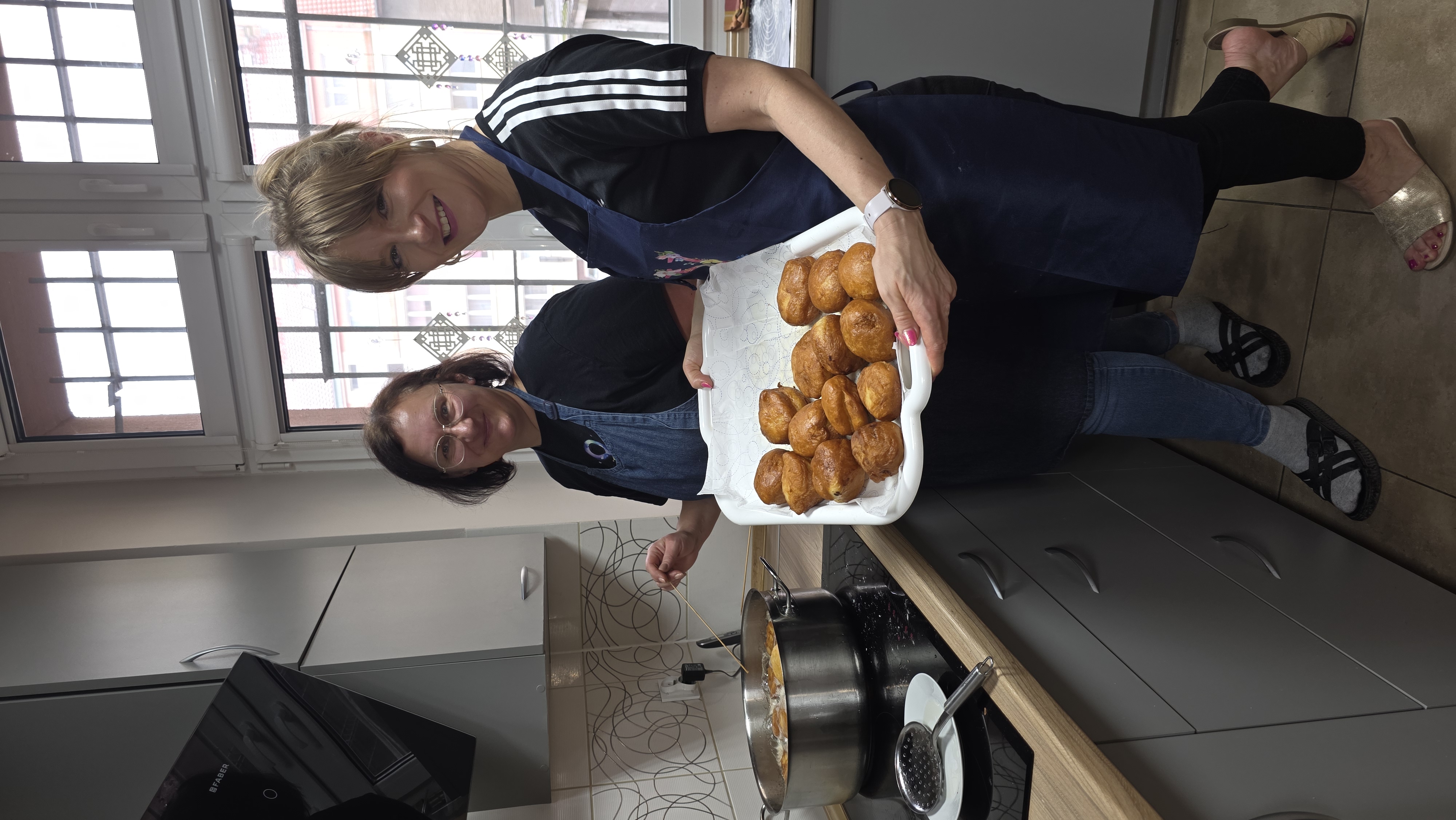 Dwie panie stoją przy garnku z tłuszczem i trzymają tacę pełną pączków.