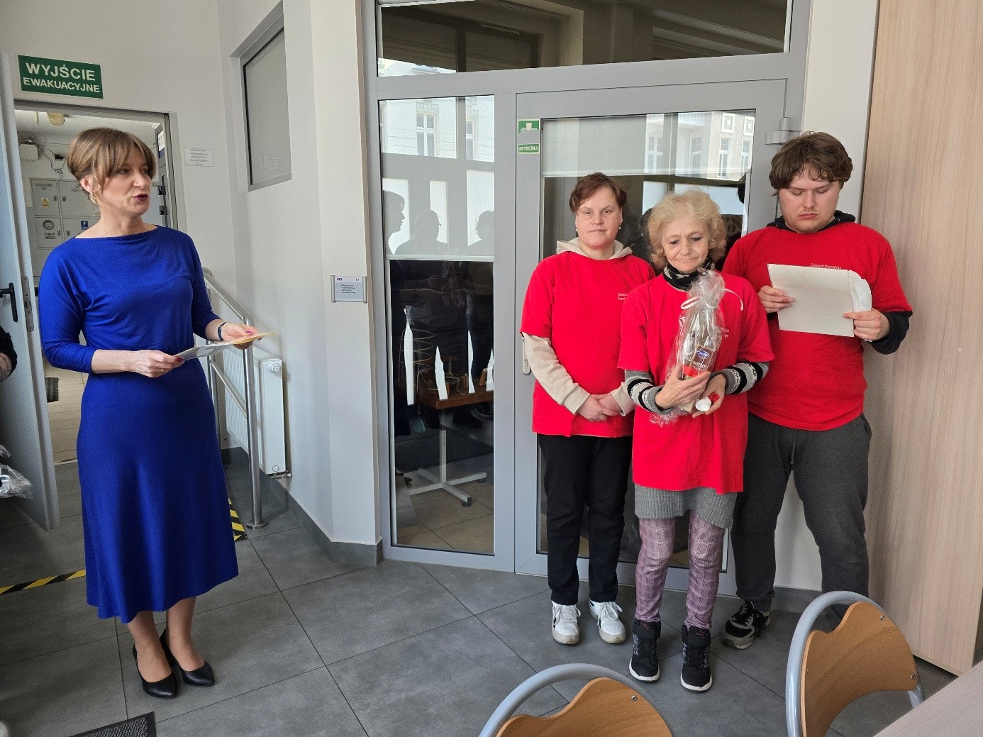 Kobieta w niebieskiej sukience, trzyma w dłoniach dyplom, który otrzymaj osoby w czerwonych koszulkach w tym dwie kobiety i jeden mężczyzna .