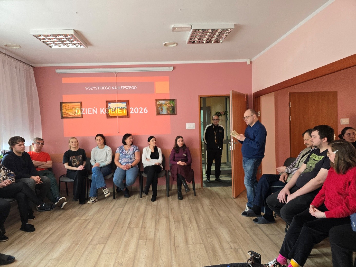 Grupa kobiet i mężczyzn siedząca na krzesłach ustawionych w okrąg. Na ścianie wyświetlony na ekranie napis Dzień kobiet.