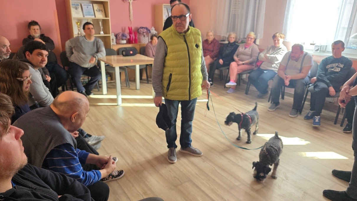 : Grupa kobiet i mężczyzn siedząca na krzesłach ustawionych w okrąg, na środku stojący mężczyzna w żółtej kamizelce trzymający w ręku dwie smycze z przypiętymi dwoma małymi psami. 