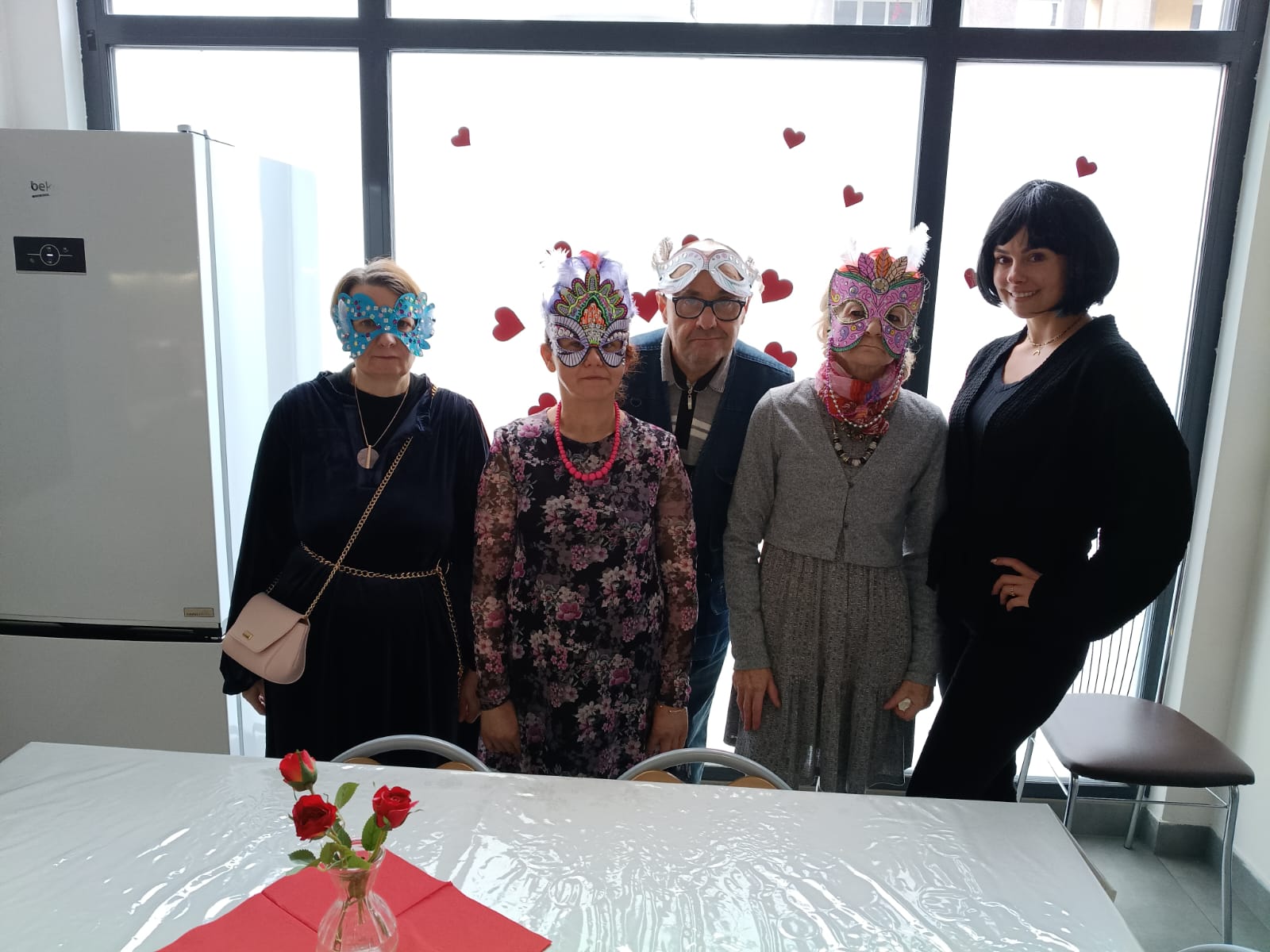 Cztery kobiety i mężczyzna stoją przy białej szybie. Cztery osoby mają maski karnawałowe na głowie.