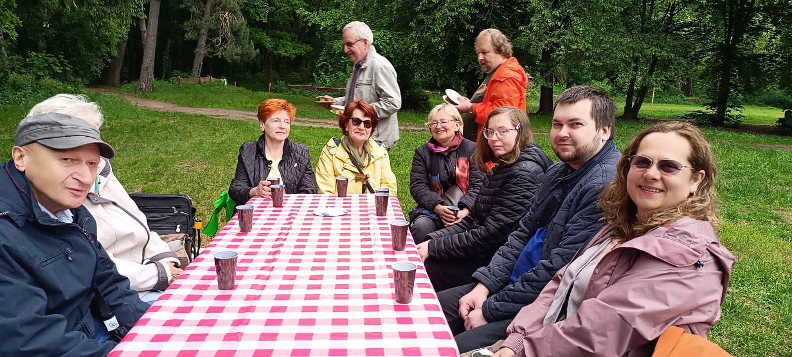 Grupa opiekunów i rodziców siedzi przy stole turystycznym na lęśnej polanie.