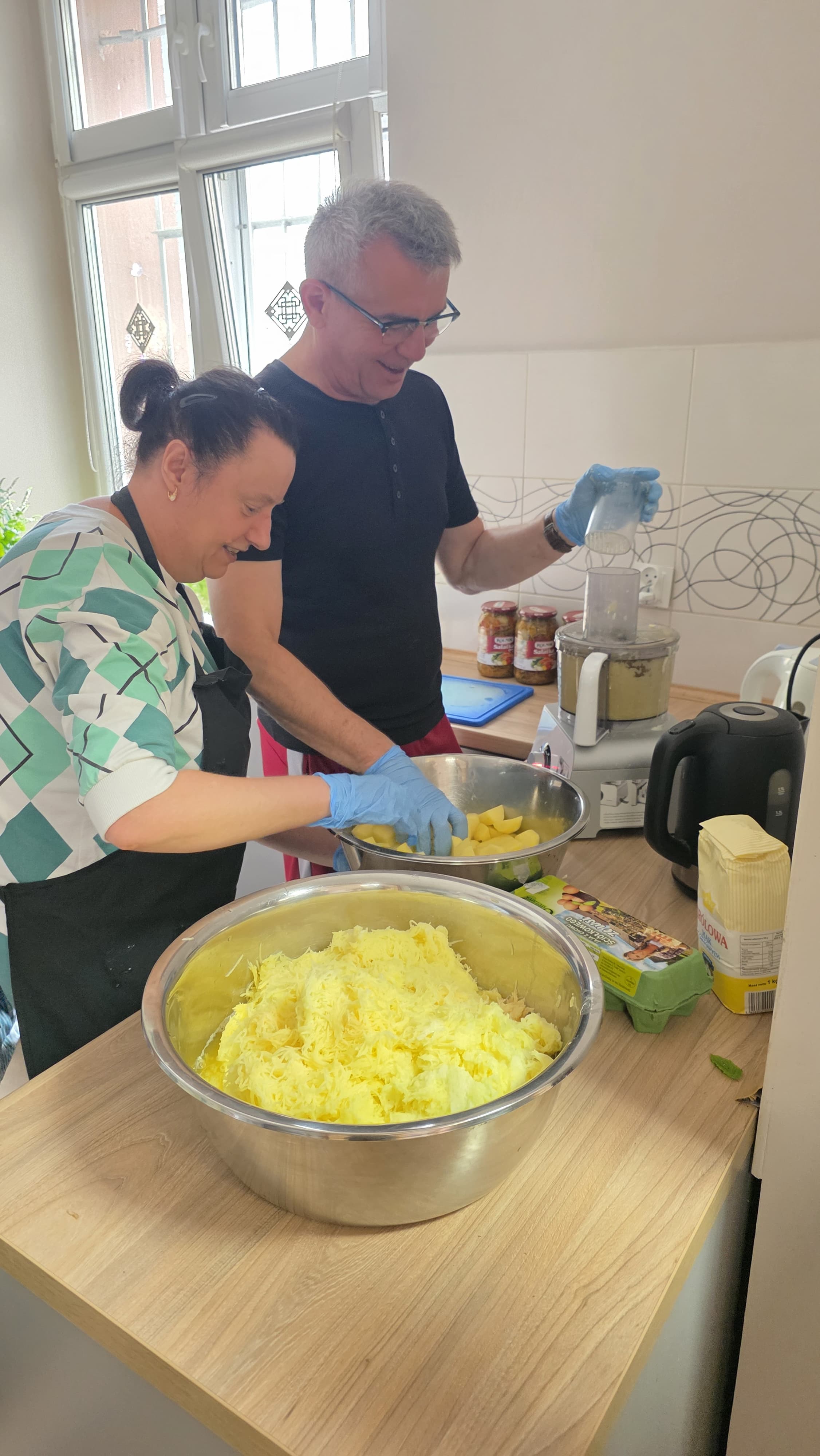 Mężczyzna i kobieta stojący przy szafkach kuchennych, na których postawianie są urządzenie do szatkowania, czajnik oraz miska wypełniona startymi ziemniakami.