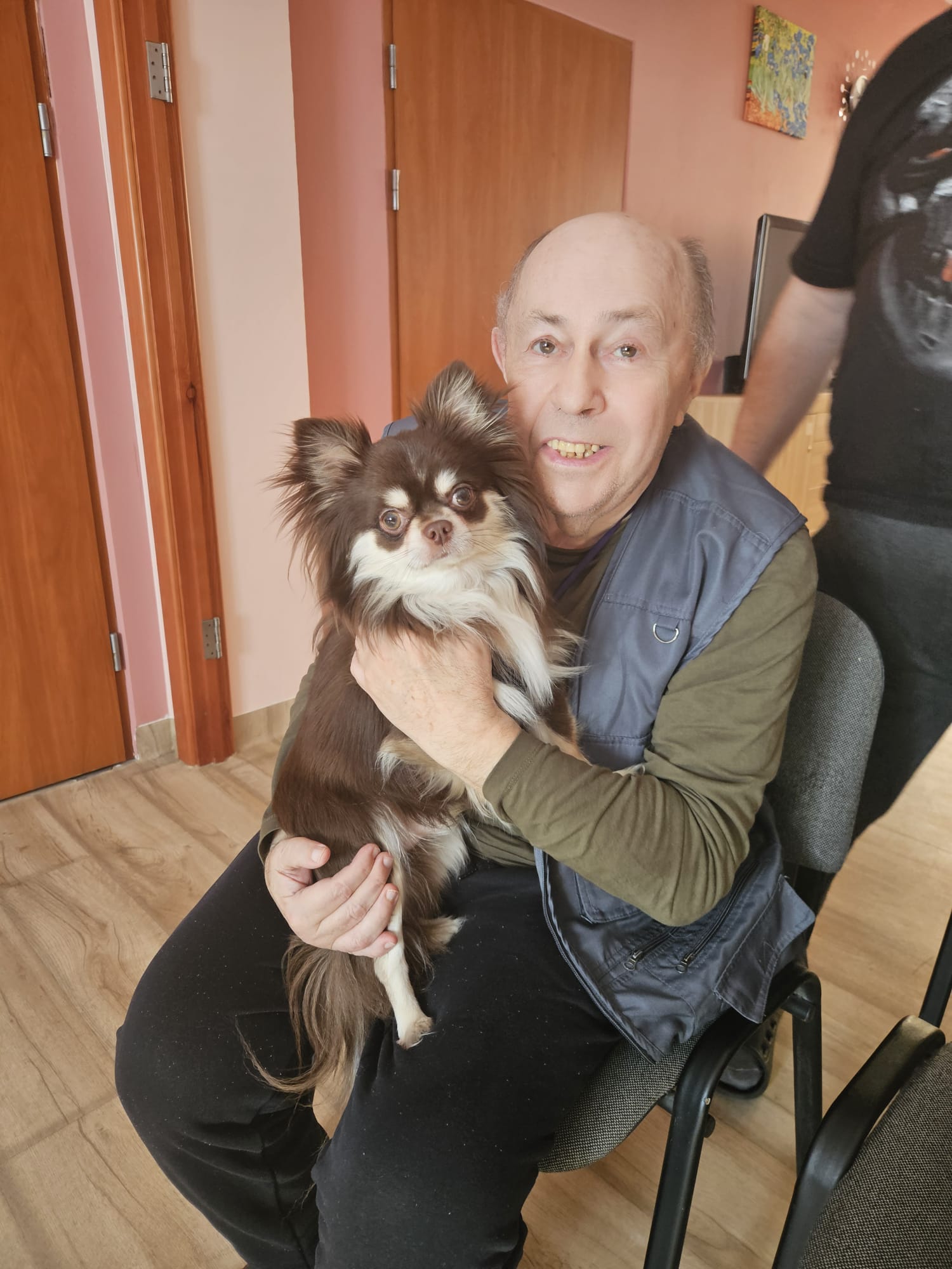 Uśmiechnięta osoba trzyma małego psa na kolanach.