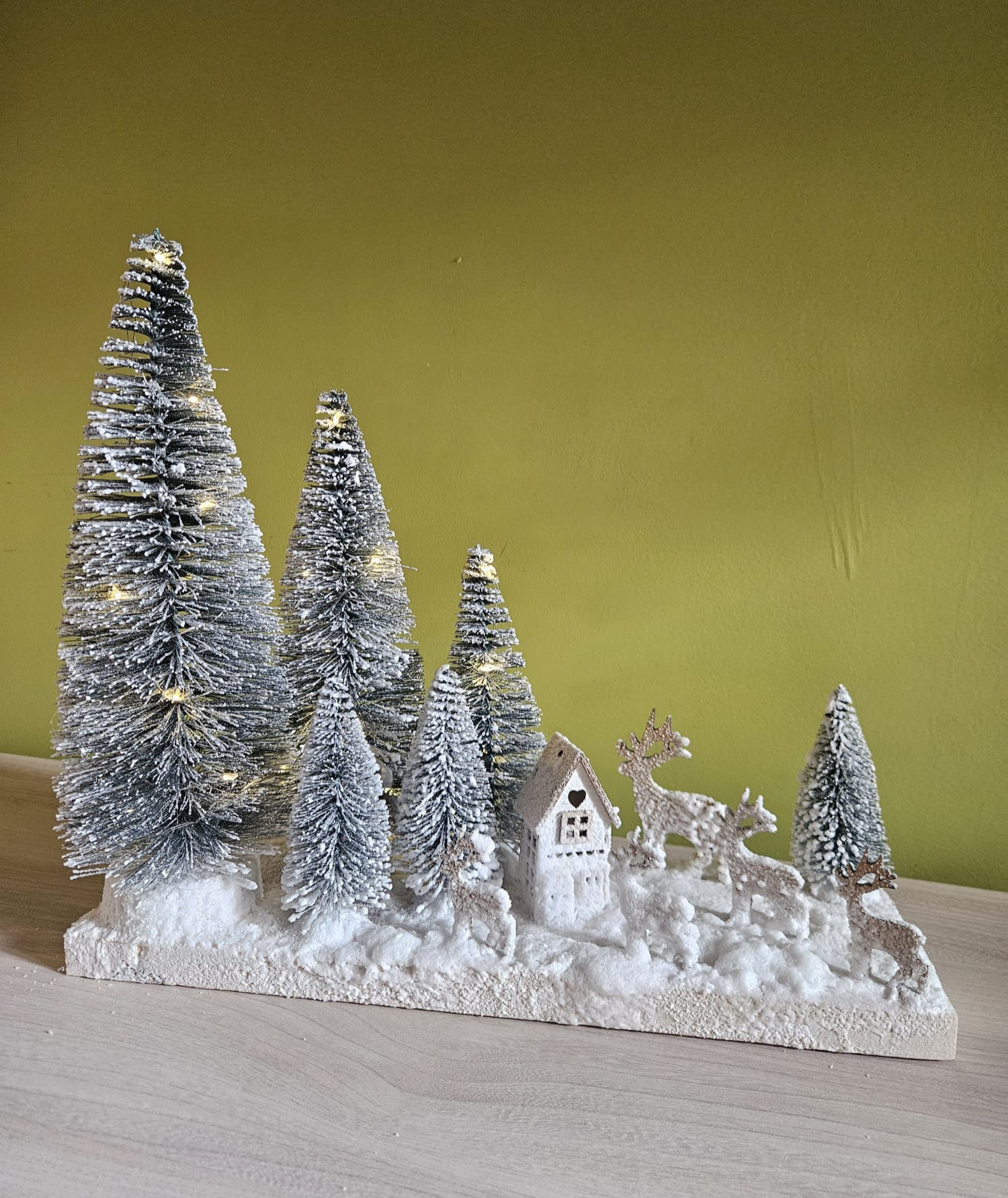 Zimowa dekoracja przedstawiająca miniaturowy zimowy krajobraz. Na białej podstawie stoją choinki, mały domek i renifery pokryte sztucznym śniegiem. Na choinkach zapalone drobne punkty świetlne.