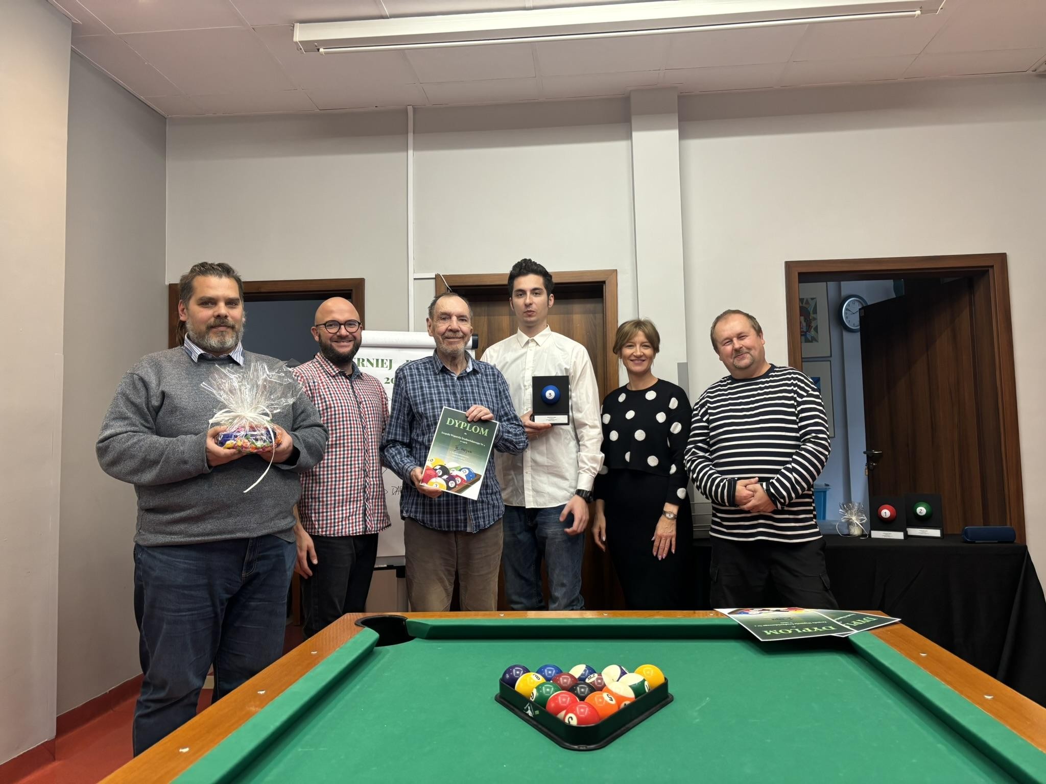 5 mężczyzn i 1 kobieta pozujący do zdjęcia. Na pierwszym planie widać stół do bilarda z kolorowymi bilami w trójkącie. Osoby na zdjęciu uśmiechają się.