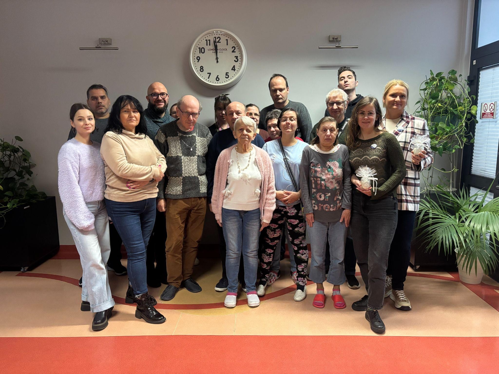 Grupa osób pozująca do zdjęcia. W tle widać duży zegar i zielone rośliny.