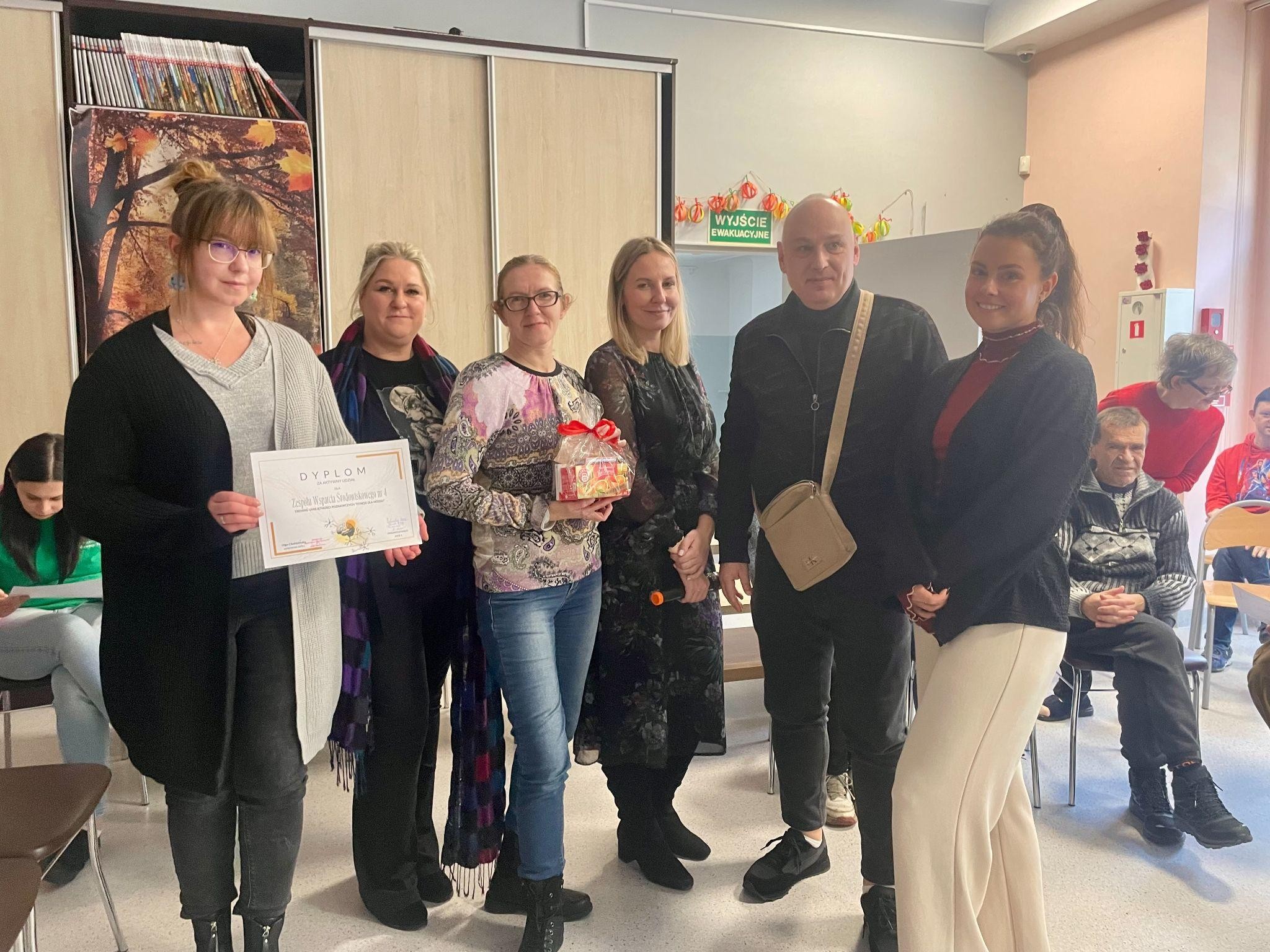 Grupa osób pozująca z dyplomem i prezentem z kokardą. W tle widać szafę, drzwi i dekoracje.