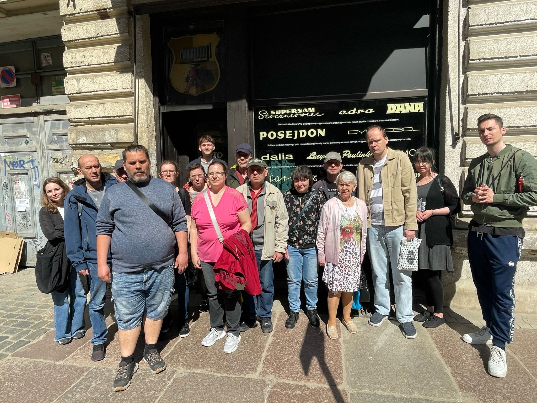Grupa osób stojąca przed witryną w kamienicy. Na szybie witryny widać napisy.