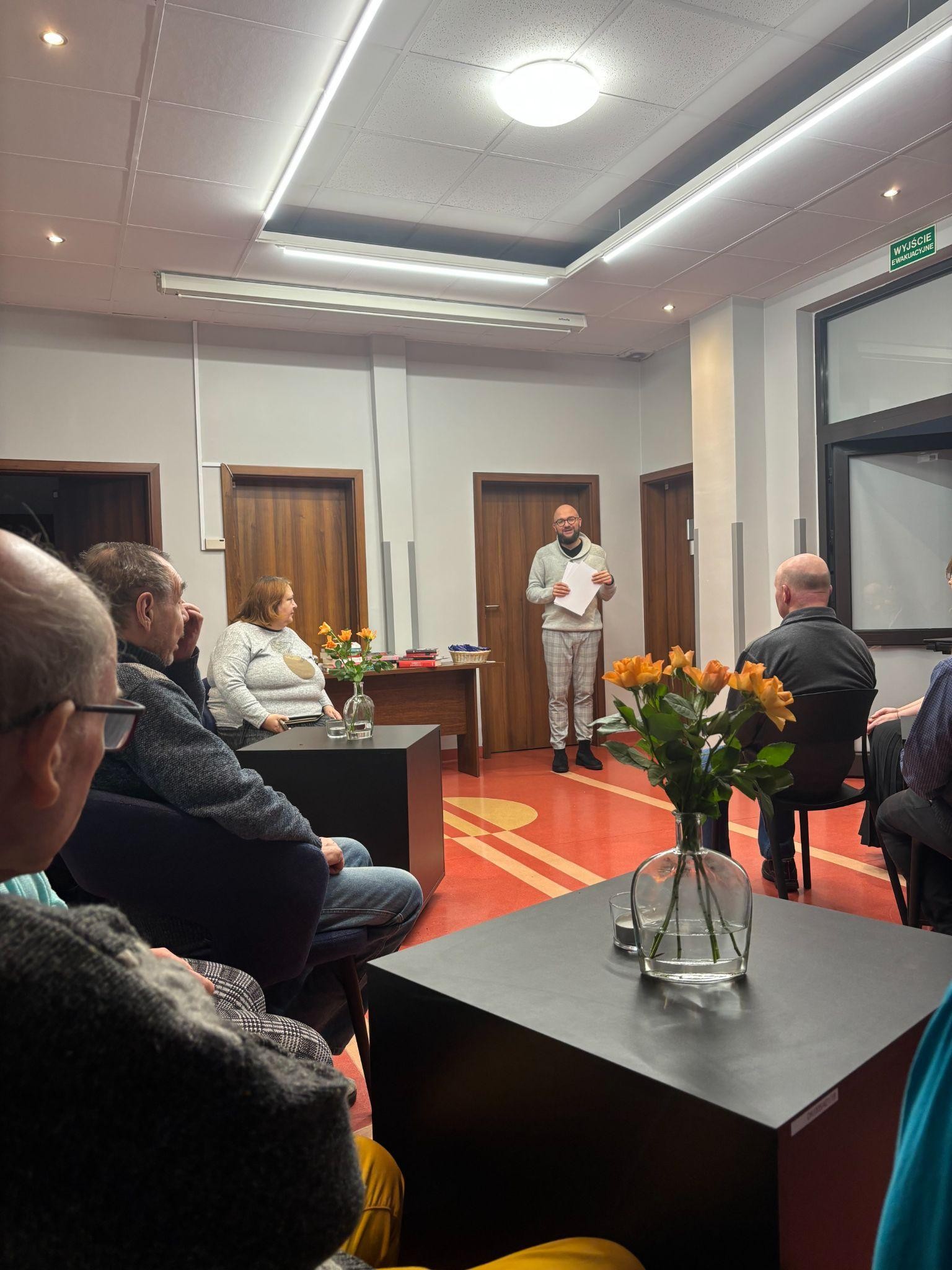 Grupa osób siedząca przy czarnych stolikach. W tle widać stojącą osobę trzymającą białe kartki oraz brązowe drzwi.