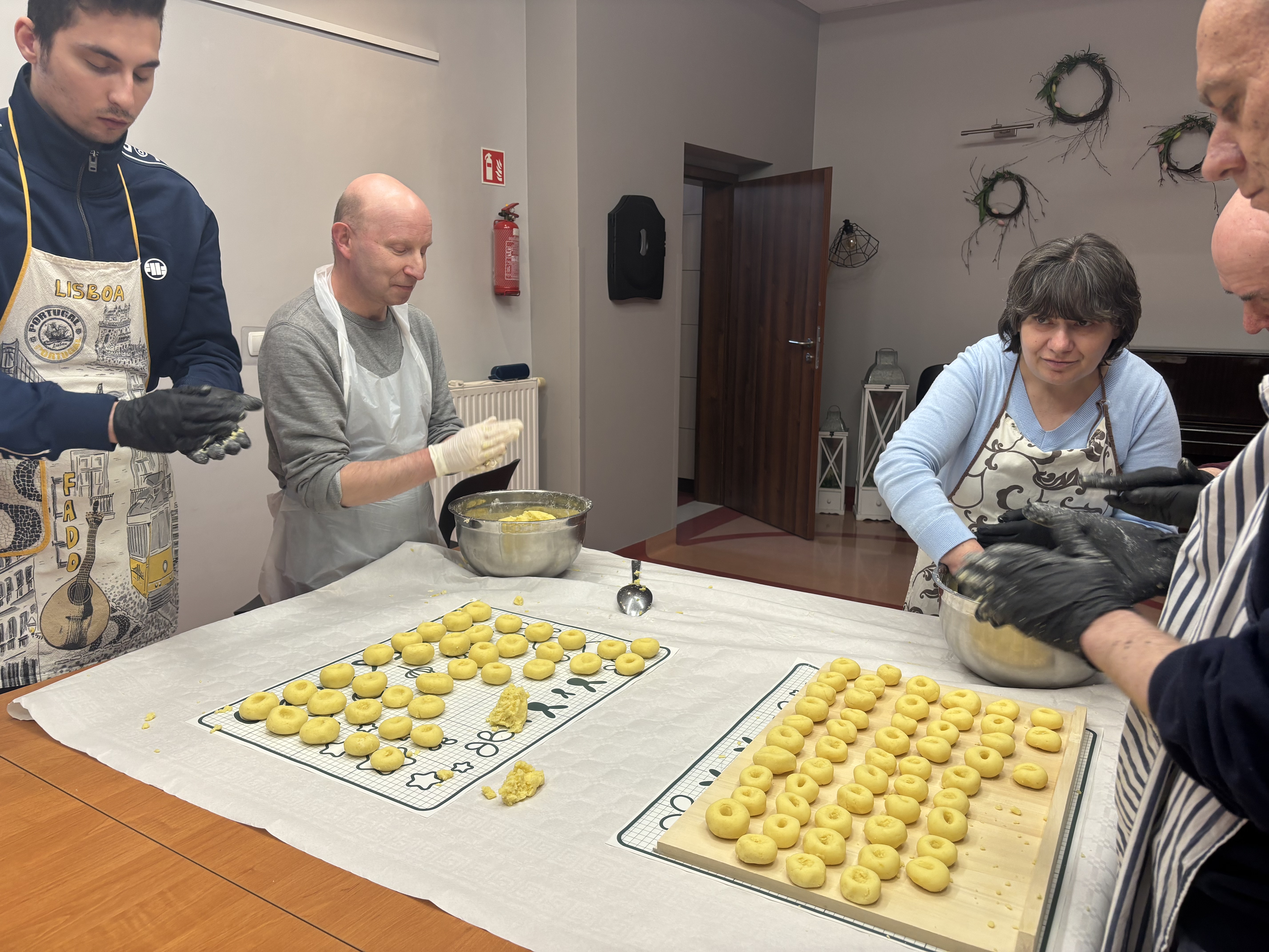 Na zdjęciu widać pięć osób. Osoby mają fartuchy oraz lepią kluski. Stoją dookoła stołu, na stole widać stolnicę i matę kuchenną na której leżą uformowane już kluski.