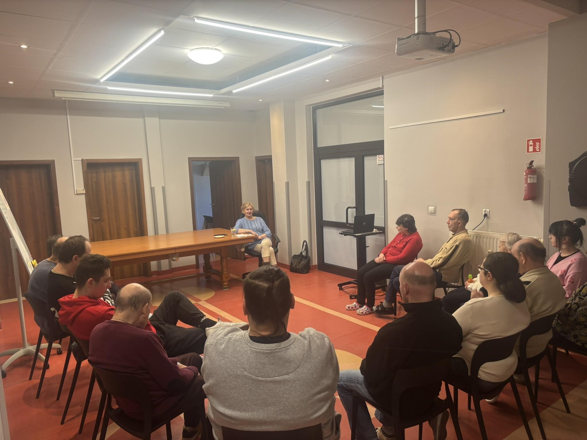 Grupa osób siedząca w kole na krzesłach. W tle widać stół, tablicę oraz drzwi.