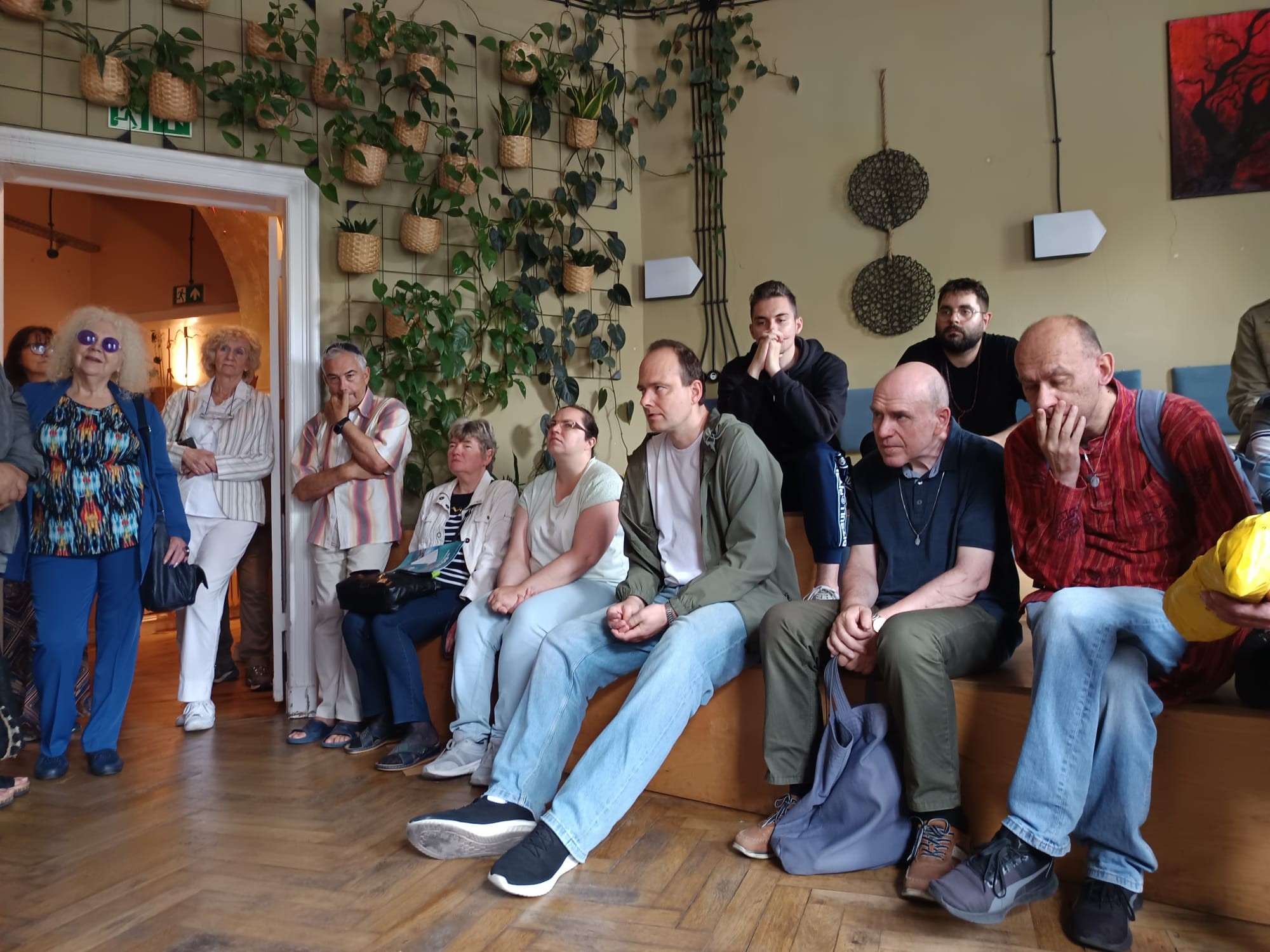 Grupa siedzących oraz stojących osób. W tle widać wiszące, zielone rośliny i dekoracje ścienne. 