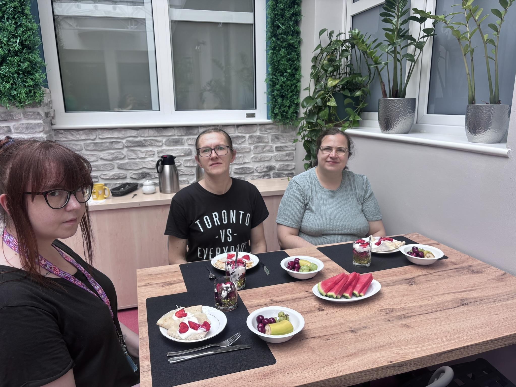 Na zdjęciu widać trzy kobiety siedzące przy stole. Kobiety uśmiechają się. Na stole widać trzy czarne podkładki. Na stole widać talerze i miski z owocami. W tle widać okna i zielone rośliny.