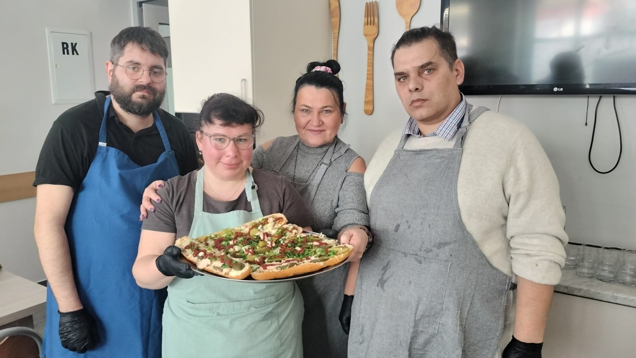 Cztery osoby w fartuchach kuchennych. Dwie osoby trzymają jedną, dużą tace. Na tacy leżą zapiekanki.