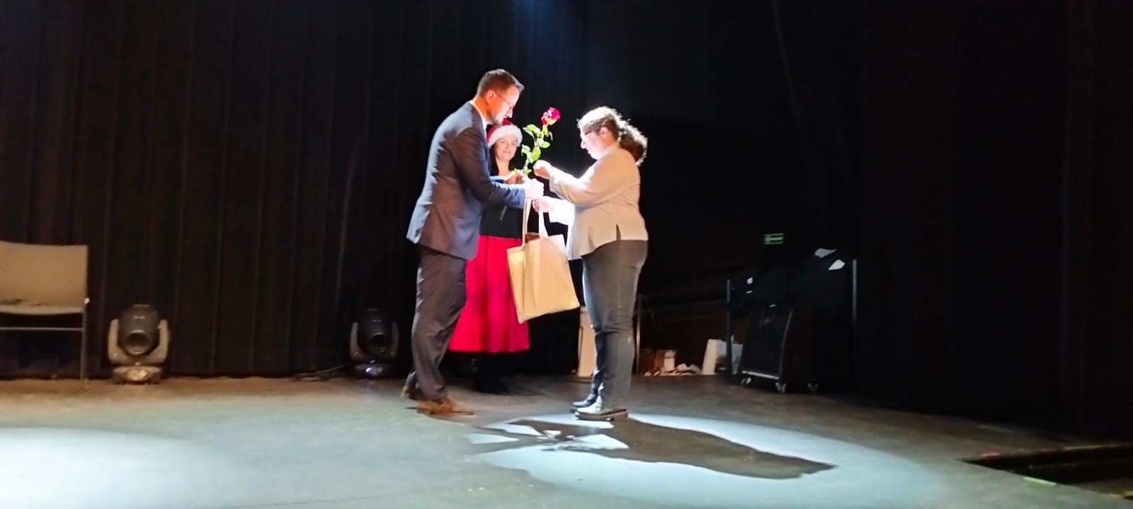 Trzy osoby na scenie. Mężczyzna wręcza kobiecie czerwoną różę i jasną torbę. Trzecia osoba stoi w tle.