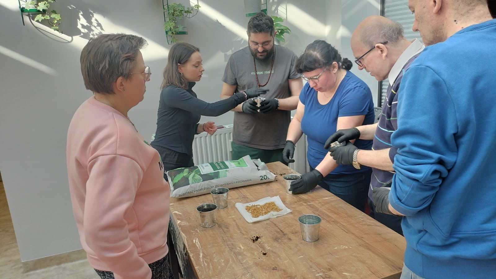 Grupa osób stojąca dookoła stołu. Osoby mają czarne rękawiczki. Na stole leży worek i stoją metalowe doniczki. W tle, na ścianie widać wiszące, zielone rośliny.