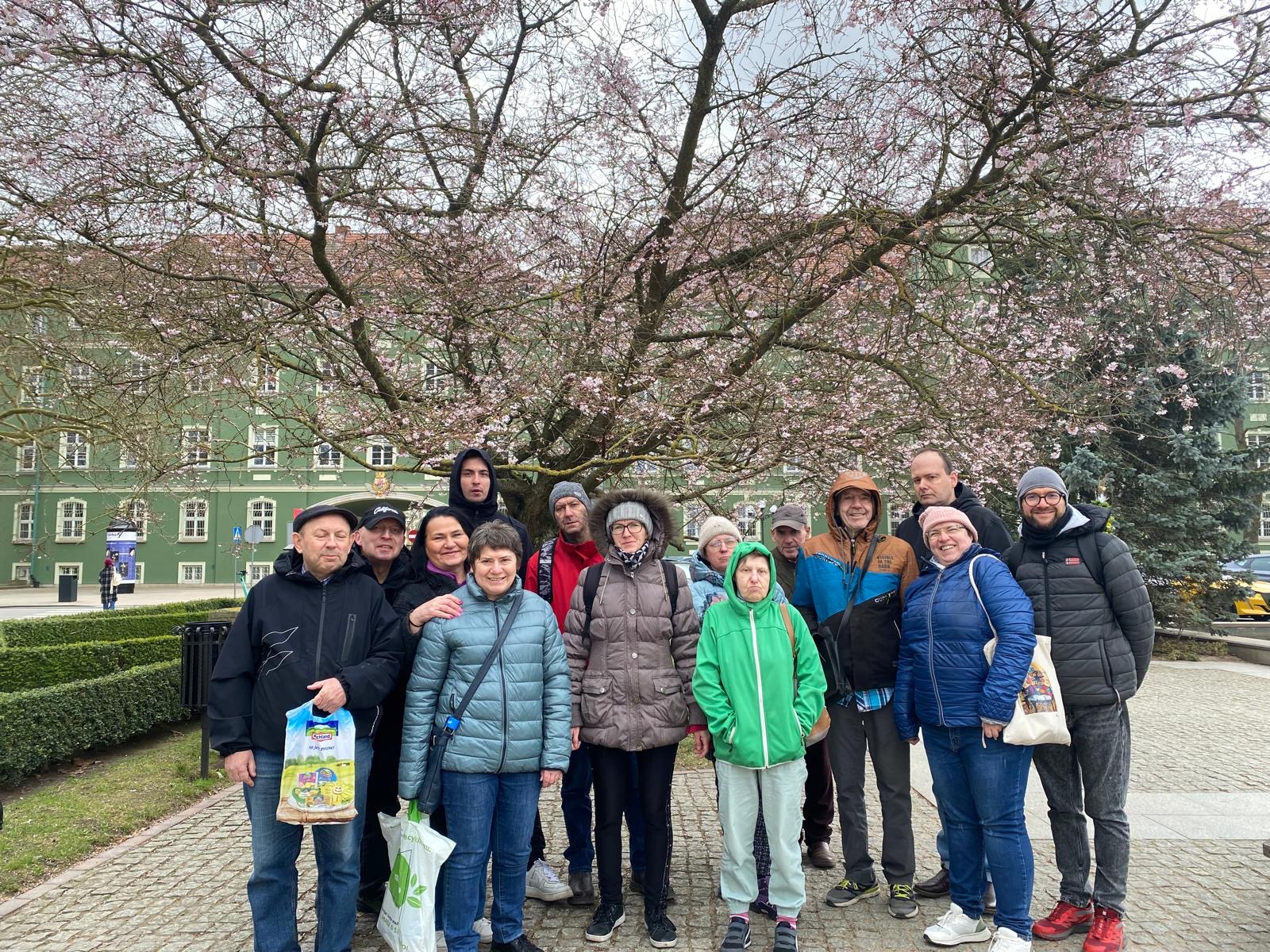 Stojąca grupa osób. W tle widać zielony budynek i drzewo z różowymi pąkami.