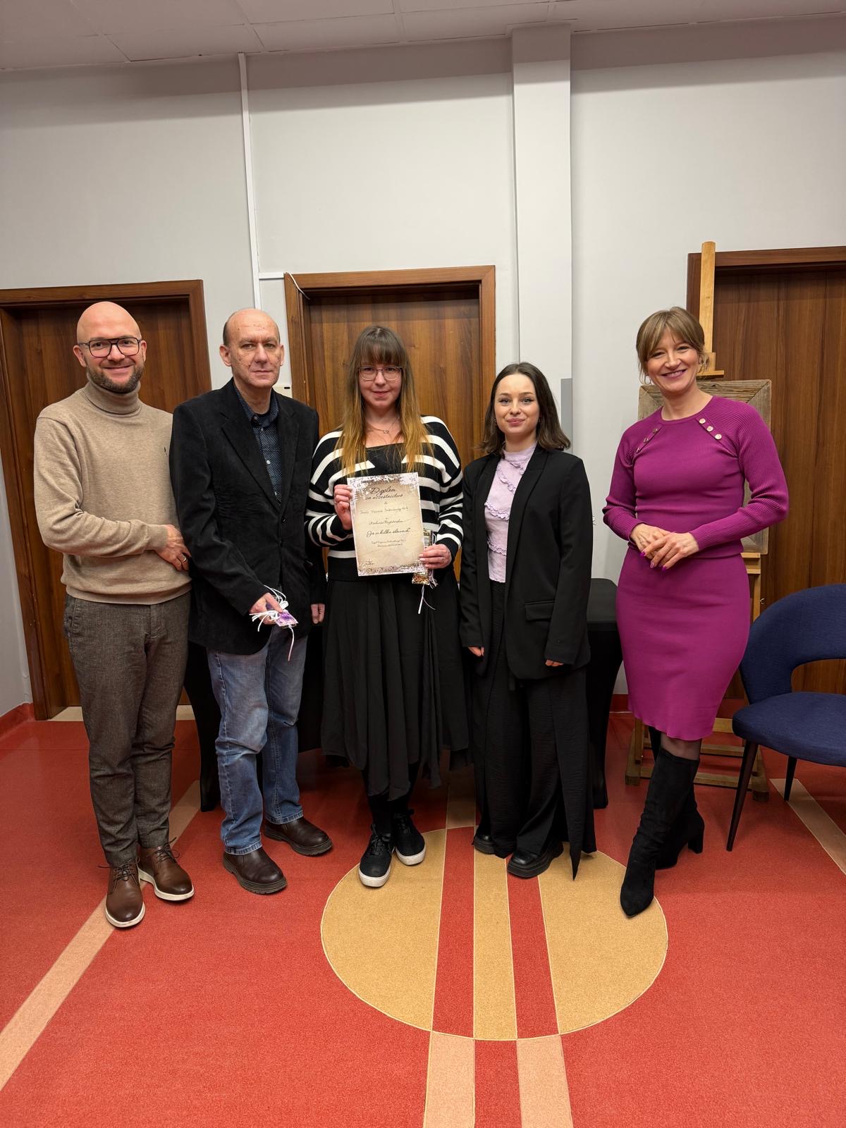 Dwóch mężczyzn oraz trzy kobiety stojące w rzędzie. Osoby się uśmiechają. Jedna kobieta trzyma kartkę. W tle widać trzy pary drzwi oraz granatowy fotel.