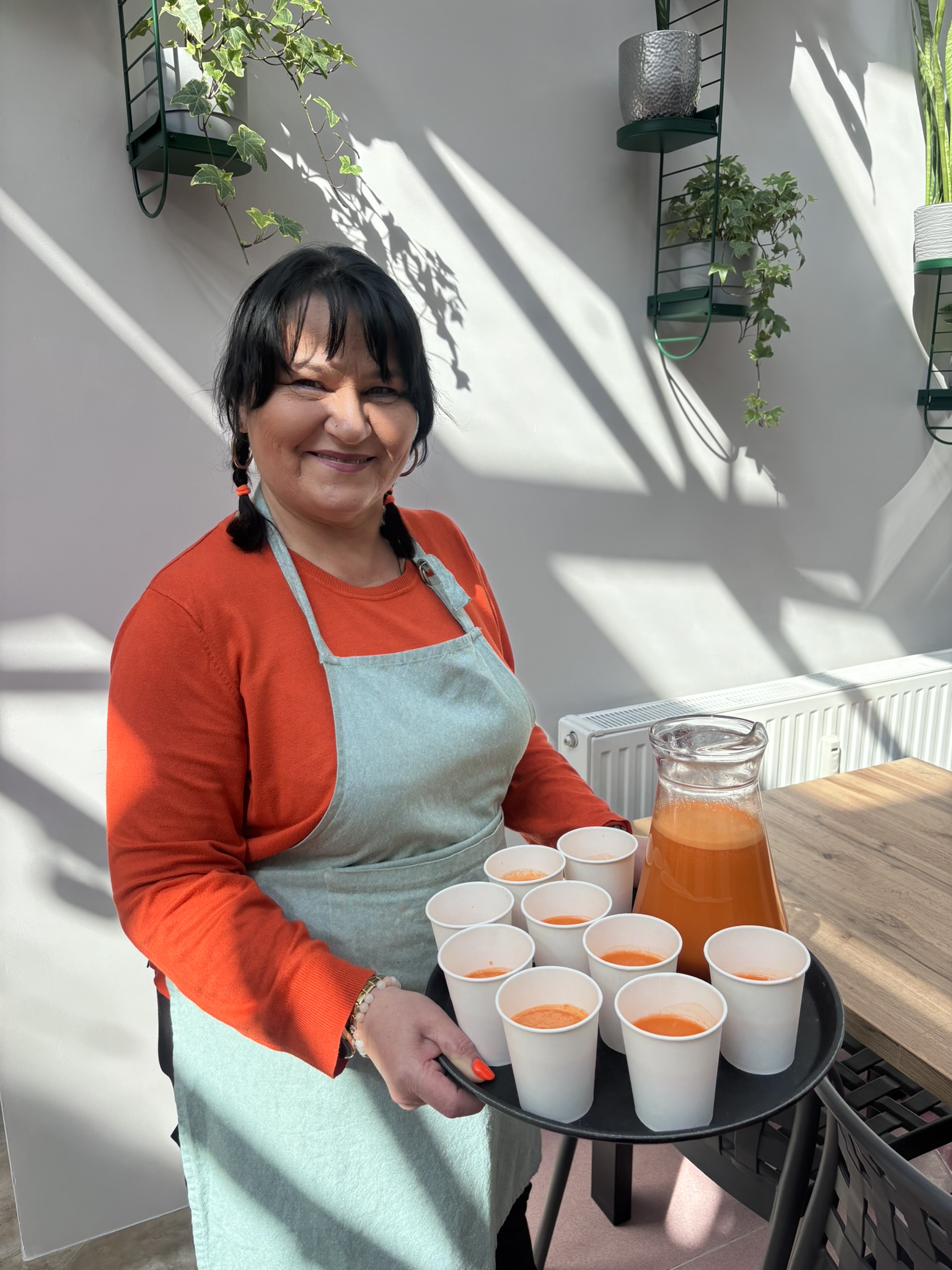 Uśmiechnięta kobieta w pomarańczowej bluzce i jasno zielonym fartuchu. Kobieta trzyma czarna tacę, na której stoją białe kubki i duży szklany dzbanek z sokiem.