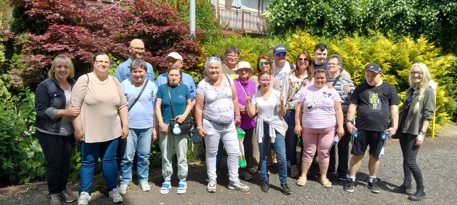 Na zdjęciu znajduje się grupa ludzi. Osoby znajdują się na zewnątrz, za nimi widać drzewa oraz krzewy.