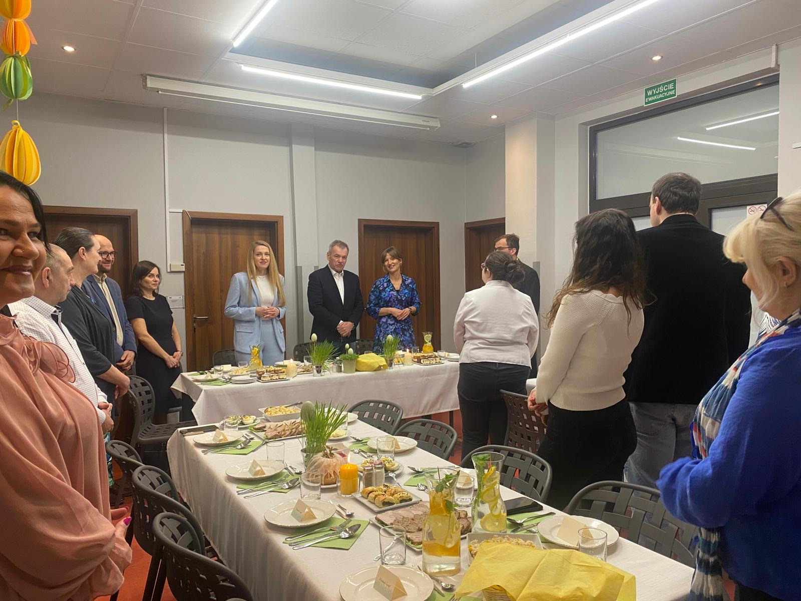 Grupa osób stojąca dookoła stołów. Na stołach leżą białe obrusy oraz biała zastawa stołowa. Na stołach widać zielone i żółte serwetki. 
