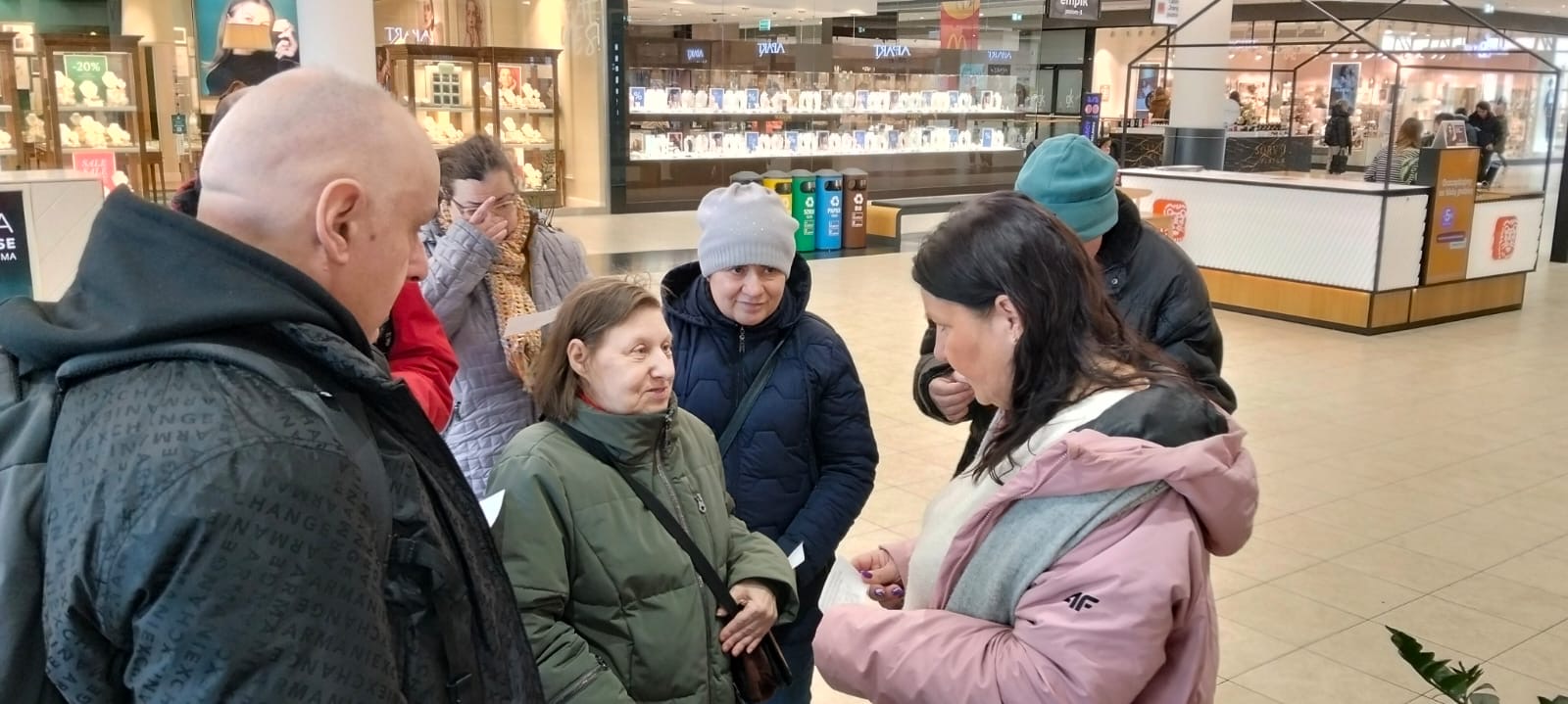 Grupa stojących osób. W tle widać sklepy oraz witryny sklepowe. 