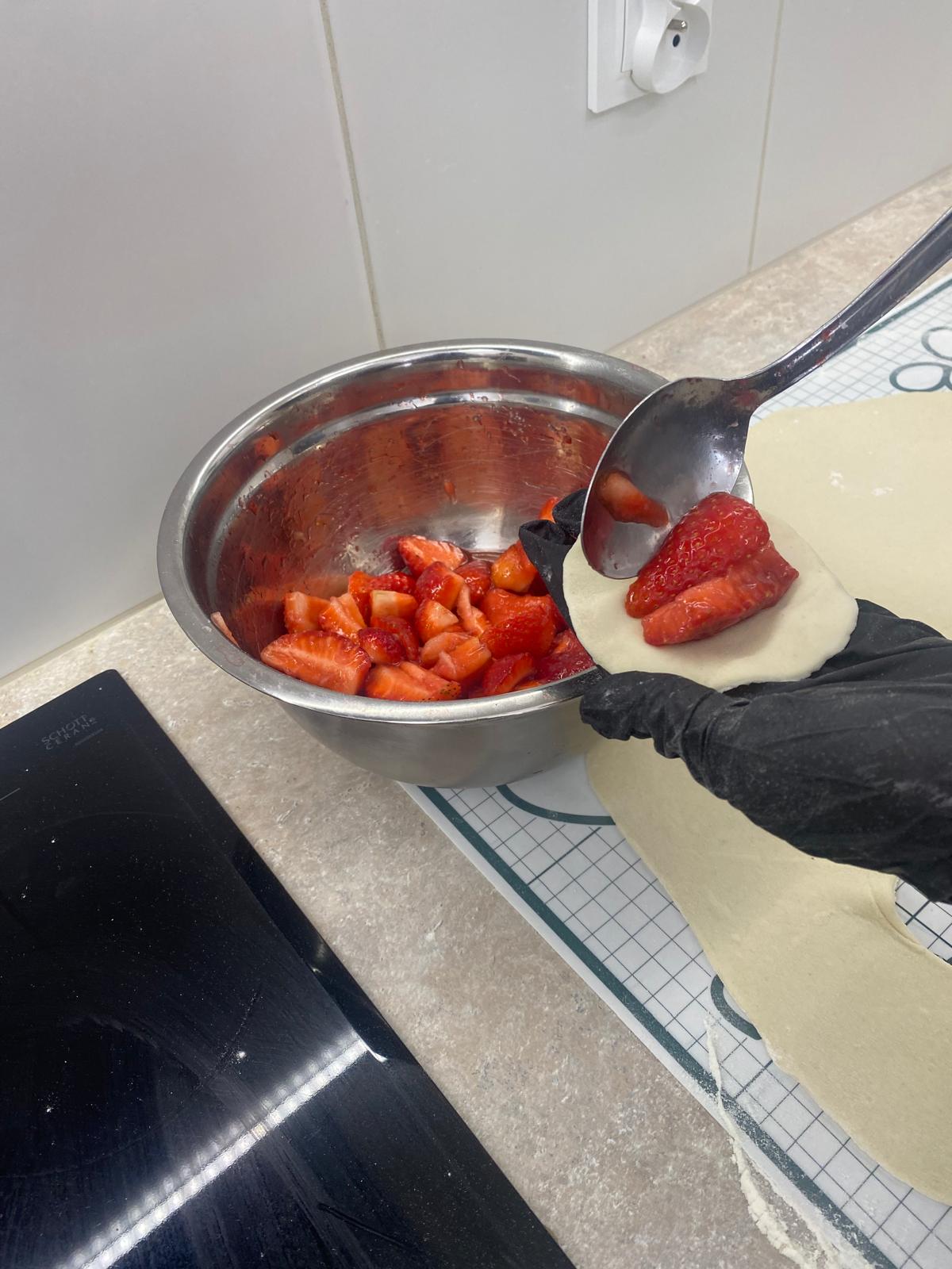 Na zdjęciu widać metalową miskę w której znajdują się kawałki truskawek. Osoba stojąca po prawej stronie nakłada truskawki łyżką na okrągły kawałek ciasta.