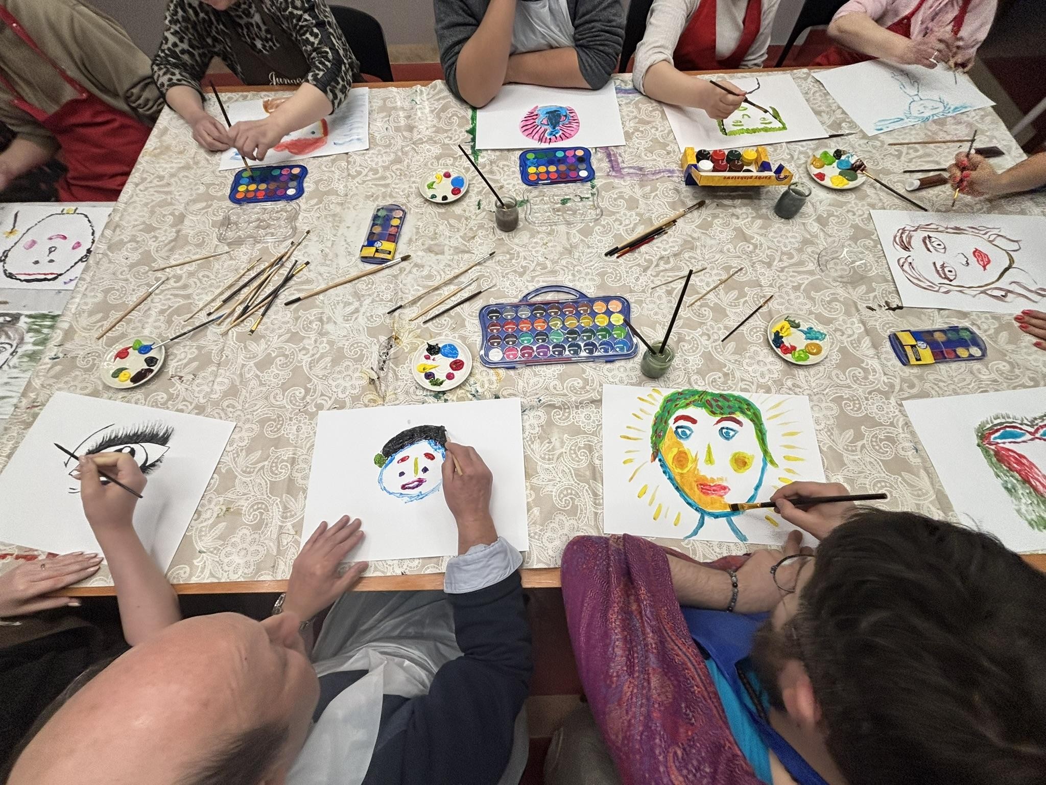 Grupa osób siedząca dookoła stołu. Osoby malują. Na stole widać farby i pędzle.