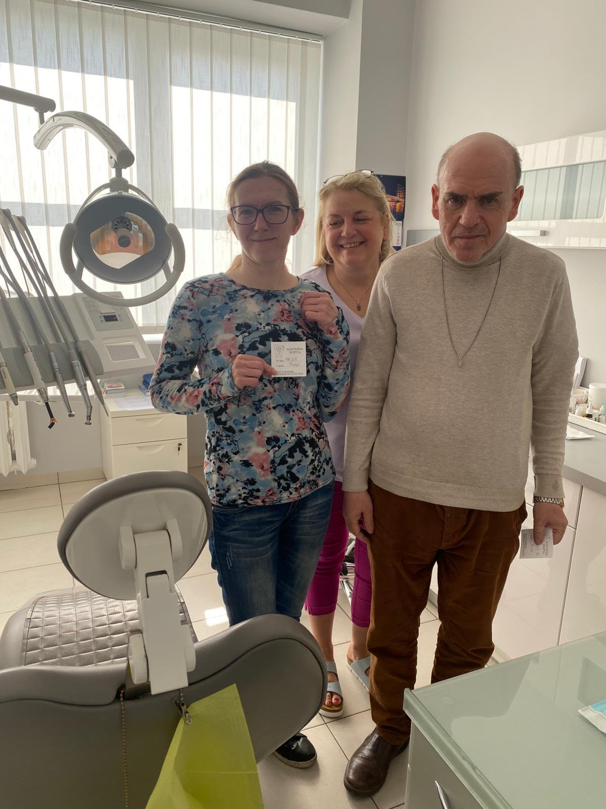 Trzy stojące osoby. Osoby się uśmiechają. Znajdują się w gabinecie stomatologicznym. Widać szary fotel,  białe szafki oraz narzędzia stomatologiczne.