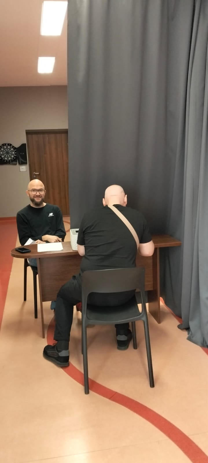 Dwóch mężczyzn siedzących na czarnych krzesłach przy stoliku z kartkami, telefonem i pojemnikiem. W tle widać zasłonę, drzwi oraz tablicę do rzutu lotkami.