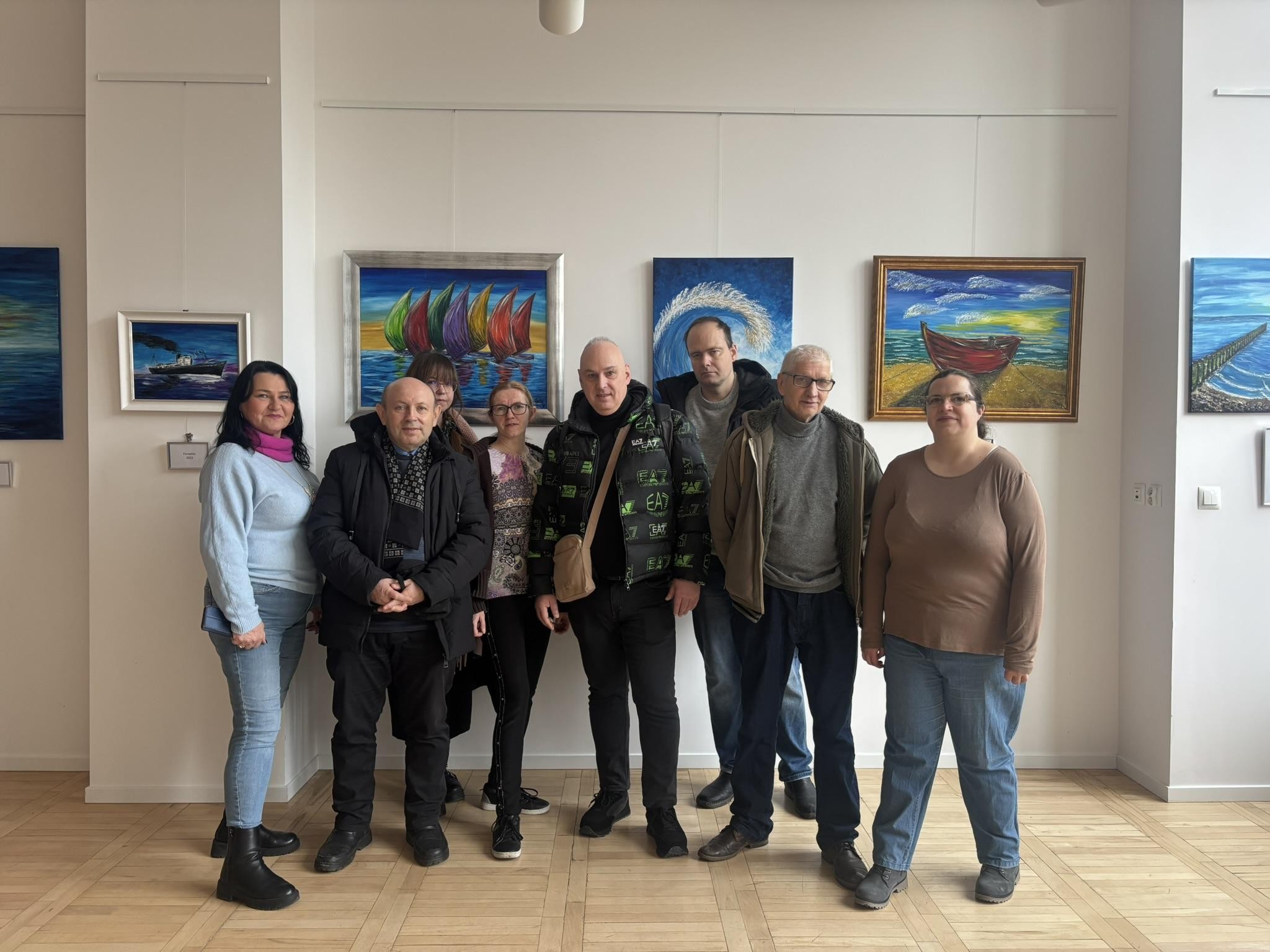 Osiem stojących osób. W tle widać obrazy malarskie na ścianach.