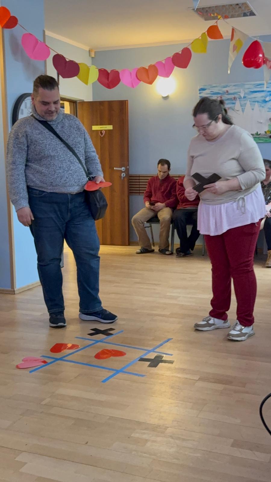 Na pierwszym planie dwie osoby. Kobieta i mężczyzna grają w grę typu kółko-krzyżyk. W tle widać osoby siedzące na krzesłach oraz dekoracje.