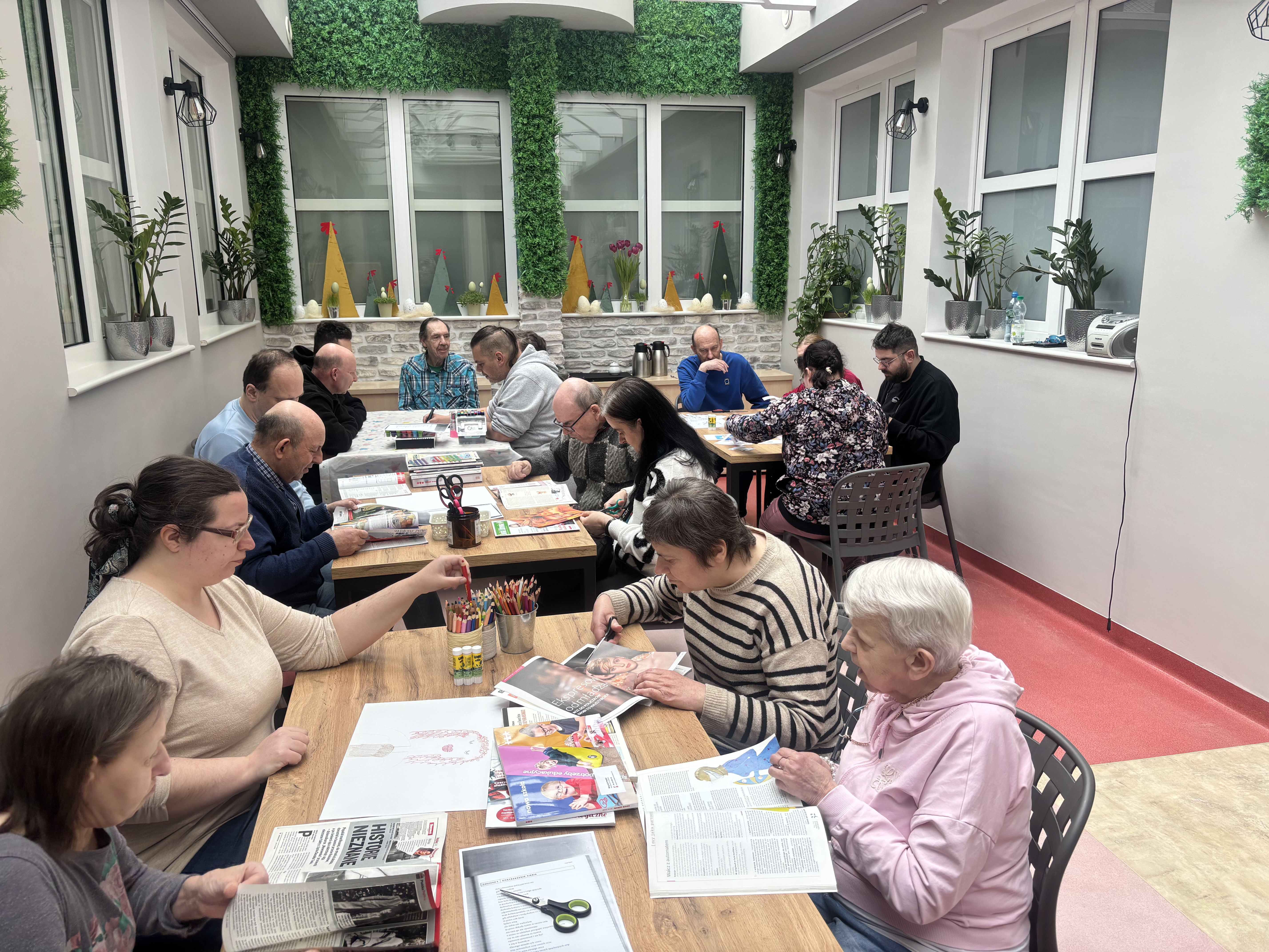 Grupa osób siedząca przy stołach. Na stołach leżą gazety, kartki, nożyczki i kredki. W tle widać okna i zieleń.