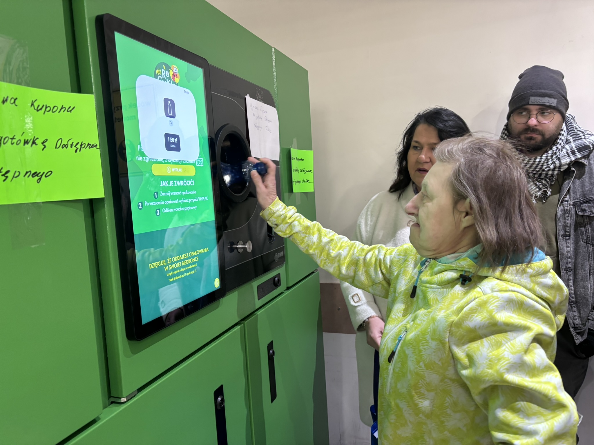 Po prawej stronie zdjęcia stoją trzy osoby. Po lewej stronie jest zielona maszyna z ekranem. Jedna z osób wkłada butelkę do maszyny.