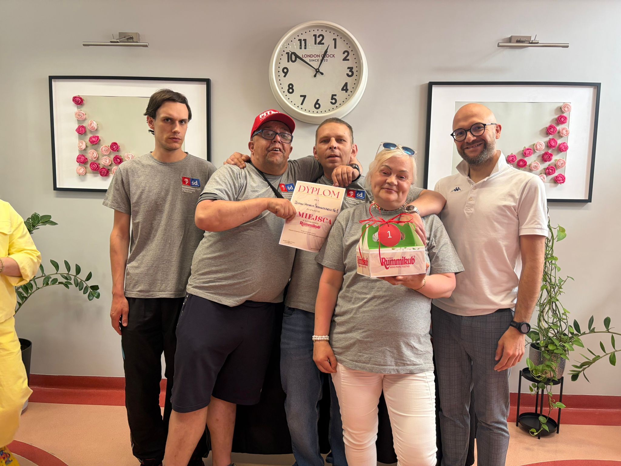 Grupa pięciu osób w szarych podkoszulkach stoi i pozuje do zdjęcia. W rękach trzymają dyplom i nagrodę w małym, pudełku. W tle okrągły zegar i dwa obrazy na ścianie.