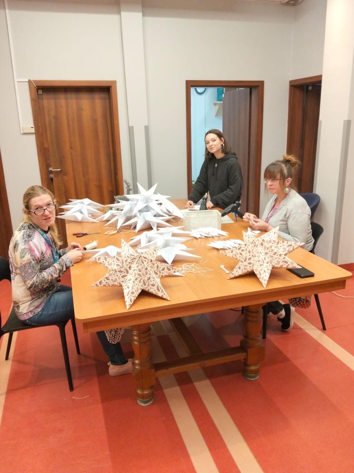 Dwie kobiety siedzące przy stole, jedna stojąca. Na stole leżą papierowe gwiazdy. W tle widać trzy pary drzwi.