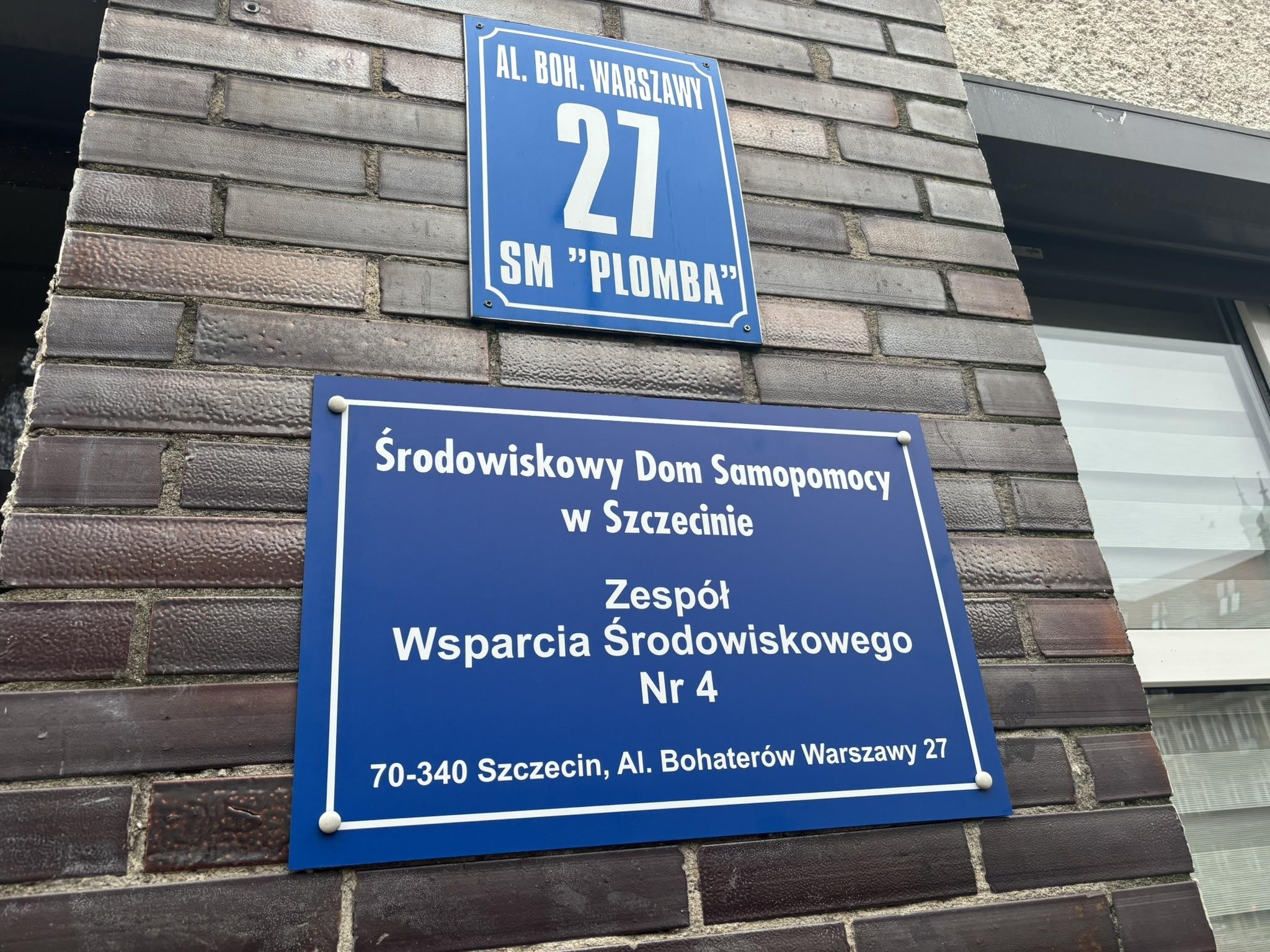 Uśmiechnięty mężczyzna stojący na zielonej drabinie. W ręce trzyma wiertarkę. Przymocowuje tablicę do zewnętrznej ściany budynku.