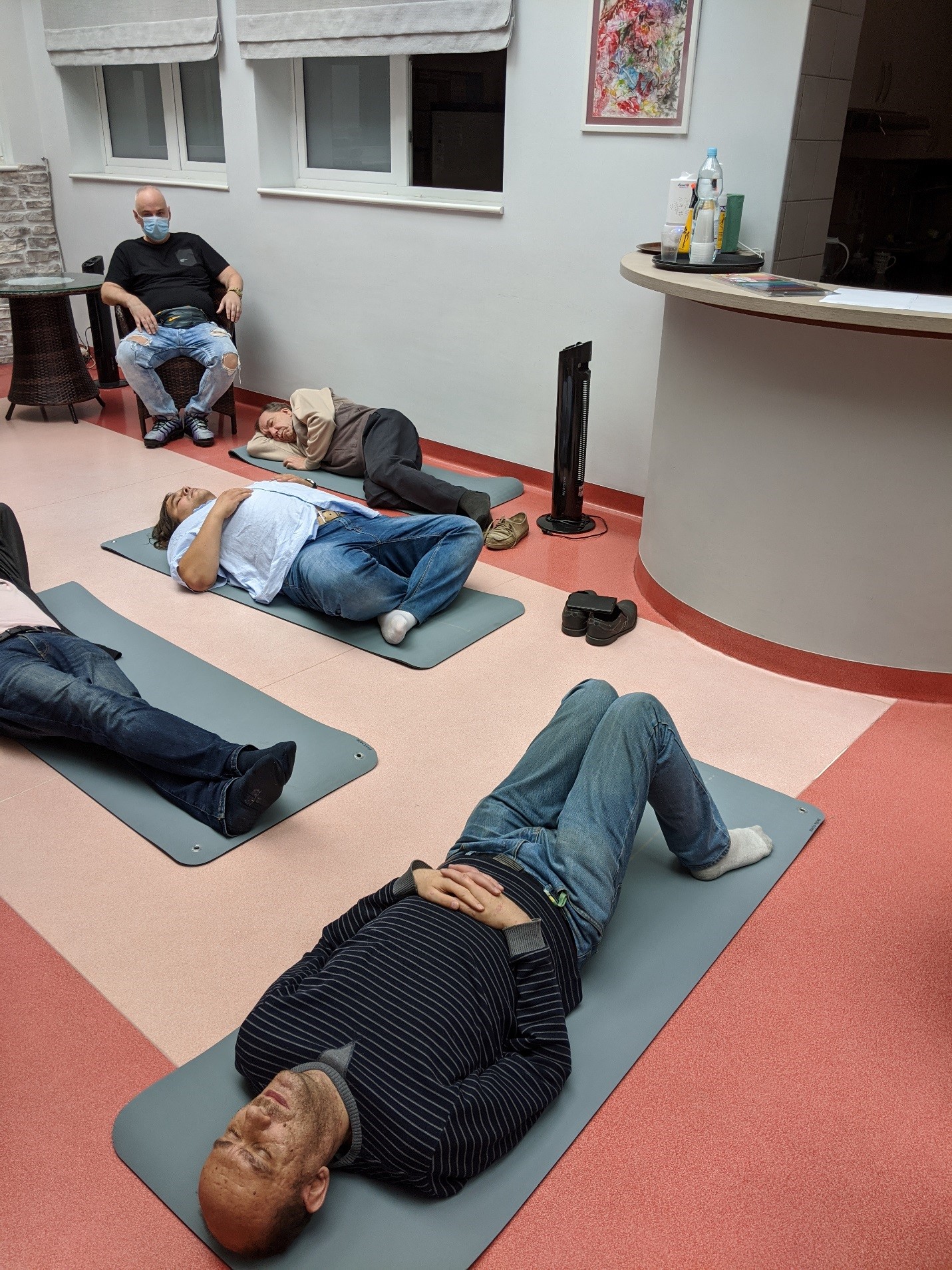 Uczestnicy terapii leżący na matach do ćwiczeń podczas zajęć relaksacyjnych, w tle mężczyzna na fotelu.