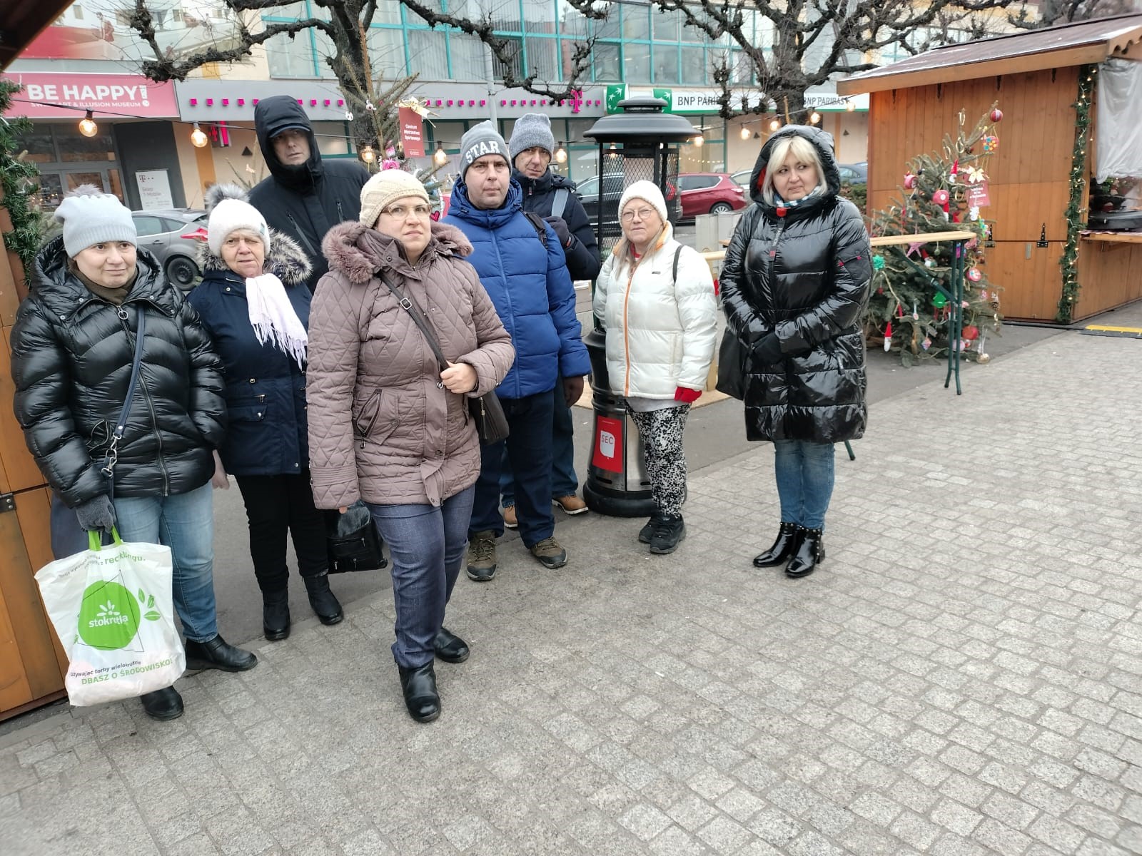 Grupa kobiet i mężczyzn na Jarmarku Bożonarodzeniowym, w tle ustrojone choinki. 