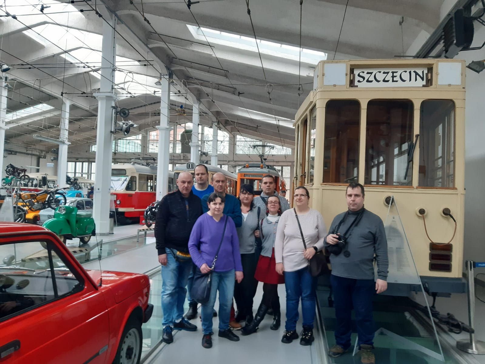 Grupa kobiet i mężczyzn w muzeum techniki i komunikacji na tle zabytkowych pojazdów. 